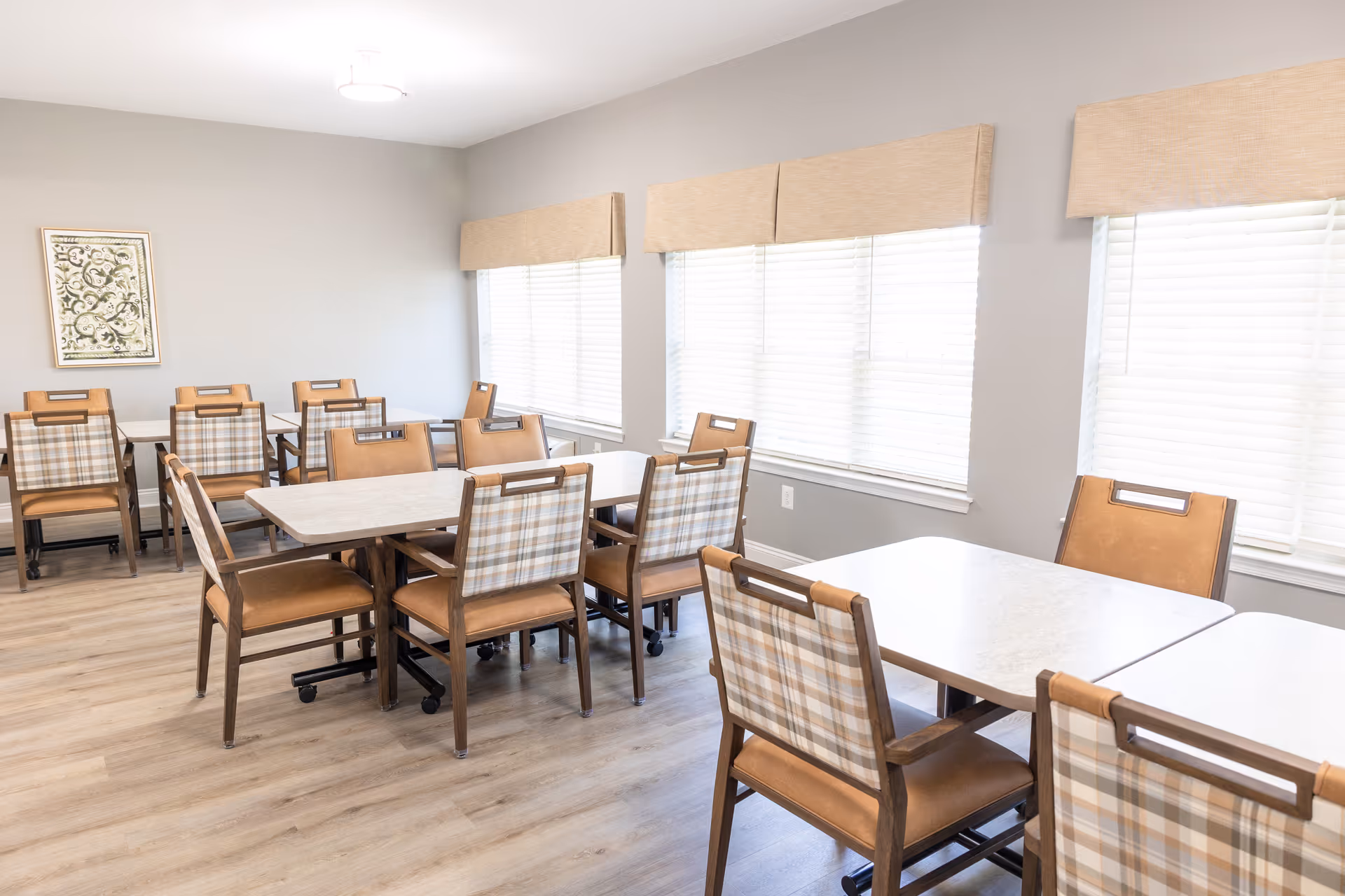 A bright dining room with multiple tables and chairs arranged neatly. The chairs have a plaid fabric backrest and brown leather seats. Large windows with beige valances let in natural light, and a framed decorative artwork hangs on the light gray wall.