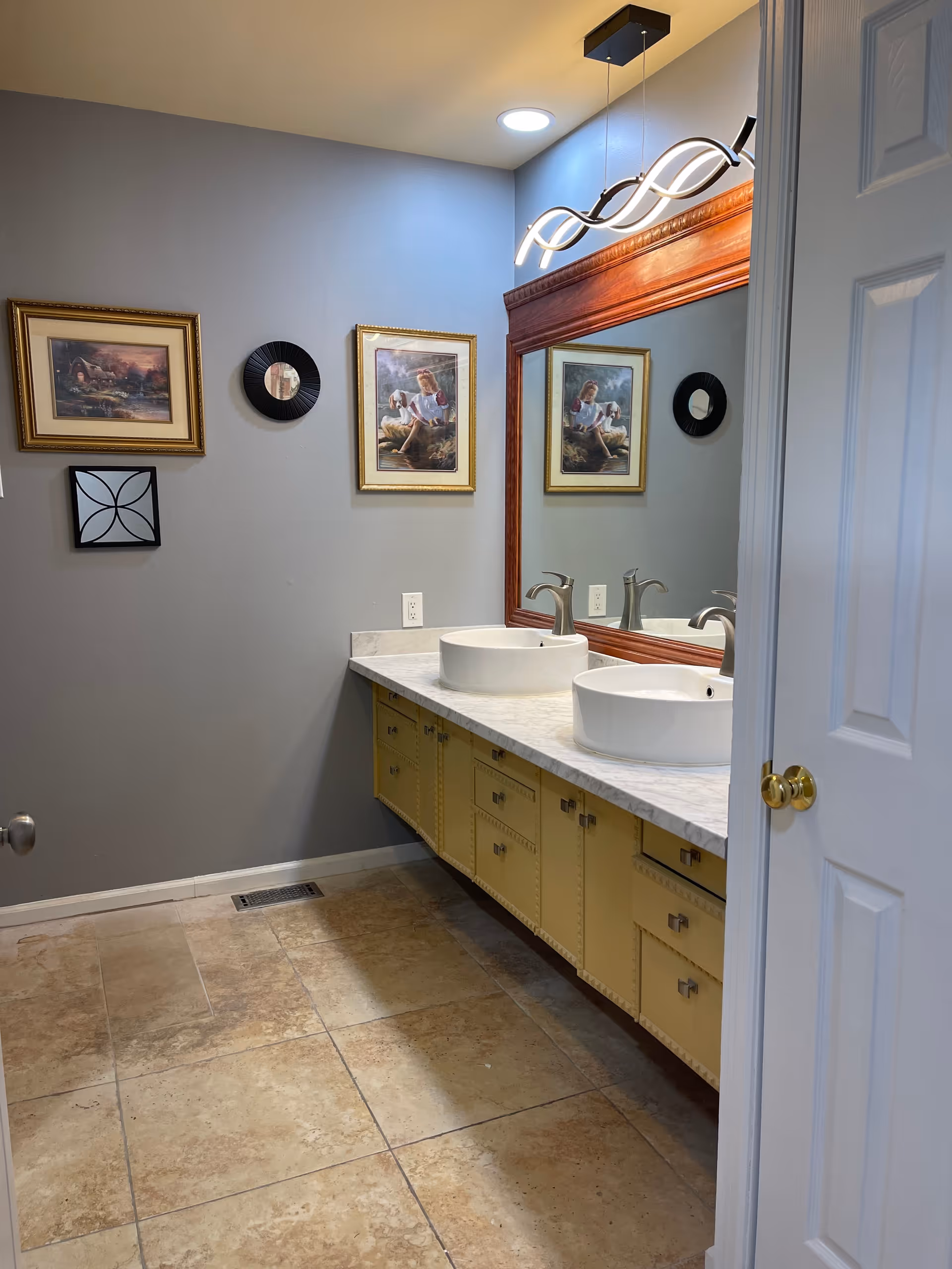 Bathroom interior with two round vessel sinks on a marble countertop, a large framed mirror, yellow under-sink cabinets, framed wall art, and tiled floor.