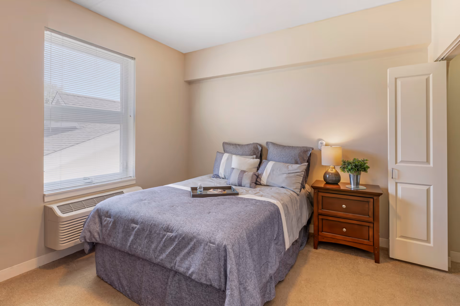 A bedroom with a neatly made bed featuring gray and white bedding and multiple pillows. There is a wooden nightstand with two drawers to the right of the bed, holding a lamp and a small potted plant. A window with blinds is on the left wall, and a partially open door is visible on the right side of the room.