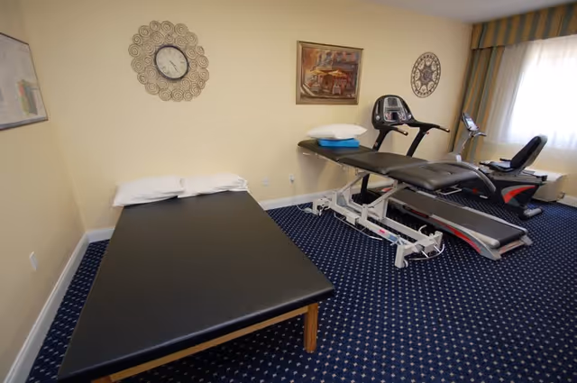 Room with a black padded treatment table with pillows, a treadmill, and an exercise bike on a blue carpet with white patterns. The walls are light yellow with a decorative clock and framed pictures. A window with striped curtains lets in natural light.