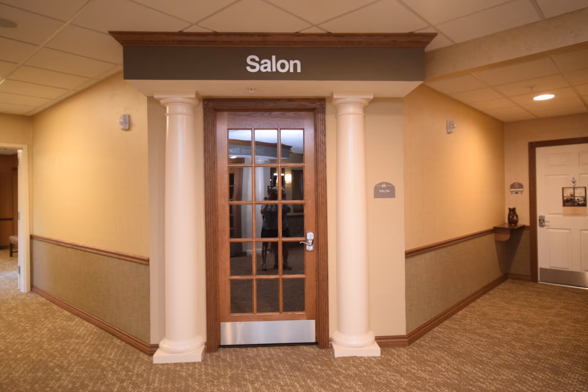 Interior hallway of Stone River Retirement Community showing a wooden door with glass panels labeled 'Salon' above it, flanked by two white columns. The hallway has beige walls with a chair rail and patterned carpet. There is a small shelf with a decorative item on the right side and another open doorway on the left.