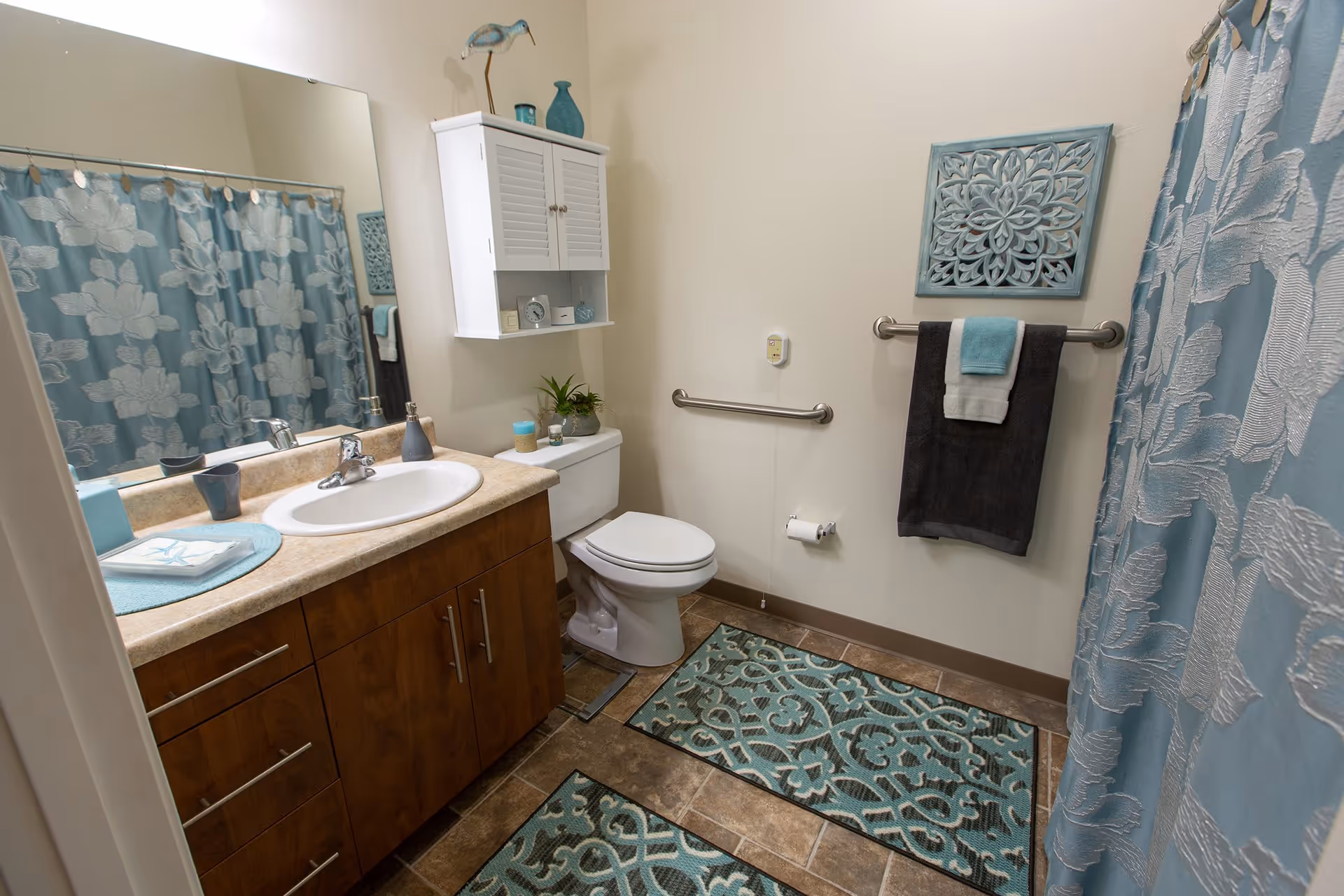 A clean and well-decorated bathroom featuring a wooden vanity with a sink and faucet, a large mirror above the vanity, a toilet with a white cabinet mounted on the wall above it, and a shower with a blue floral-patterned curtain. The bathroom has blue and brown patterned rugs on the floor, a towel rack with folded towels, and decorative items including a small plant and wall art.