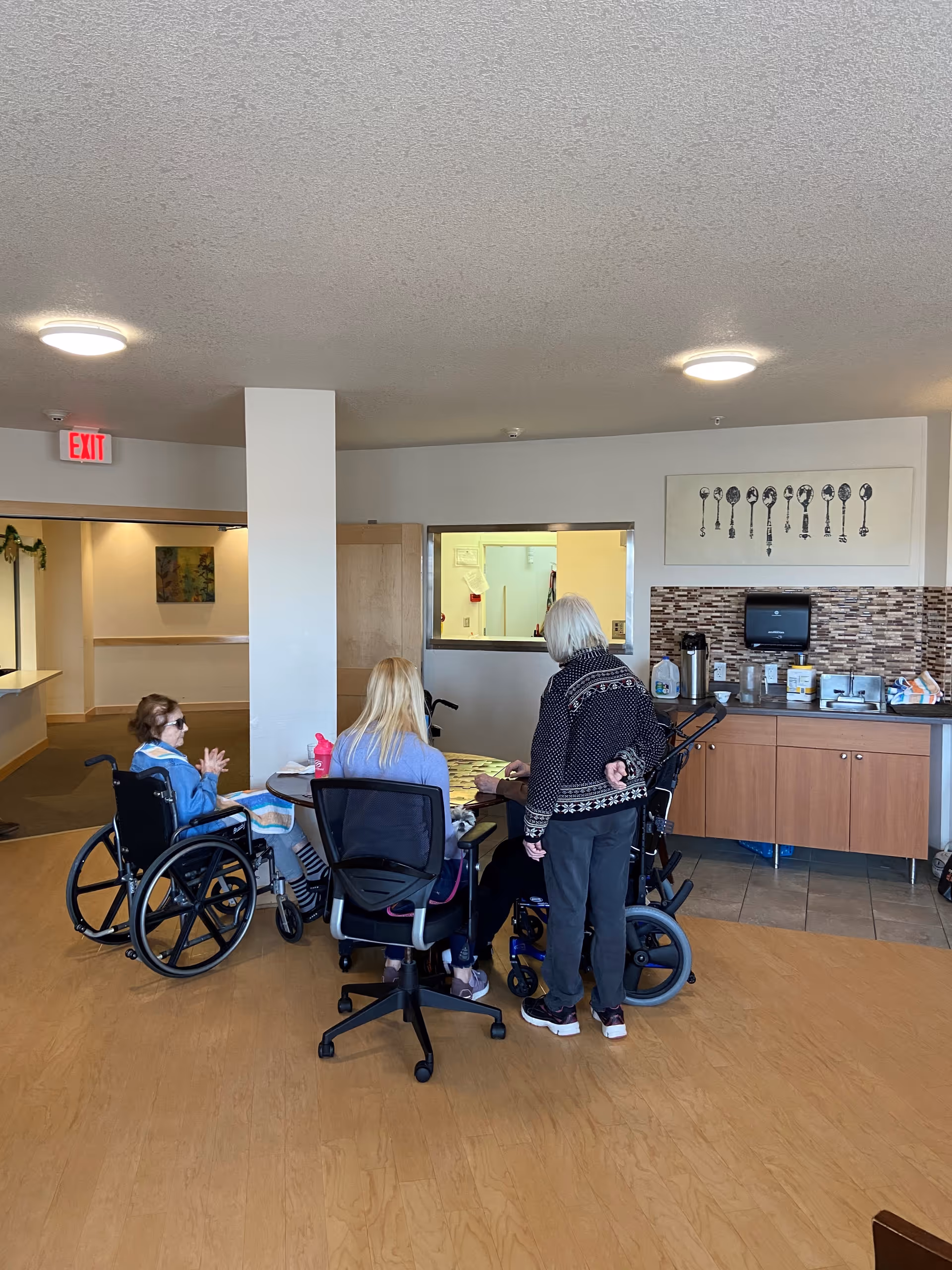 A group of elderly individuals, some in wheelchairs, and a caregiver sitting and standing around a table in a common area of a senior living facility. The room has wooden flooring, a kitchenette area with cabinets and a coffee station, and a wall decoration featuring a row of vintage keys.