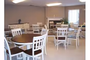 A spacious dining room with multiple round wooden tables surrounded by white chairs. The room has beige walls, a fireplace in the background, and large windows allowing natural light to enter. There is a sideboard against one wall with decorative items on top.