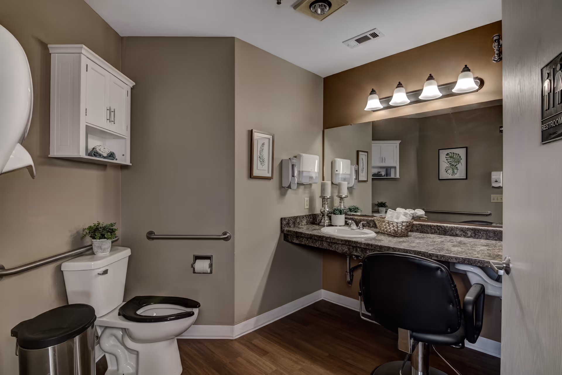 A clean and accessible bathroom with a white toilet featuring a black seat, a stainless steel trash can, and a white wall cabinet above the toilet. The bathroom has beige walls, a large mirror above a granite countertop with a sink, a black cushioned chair in front of the counter, and wall-mounted soap and paper towel dispensers. There are framed botanical prints on the walls and a handrail next to the toilet.