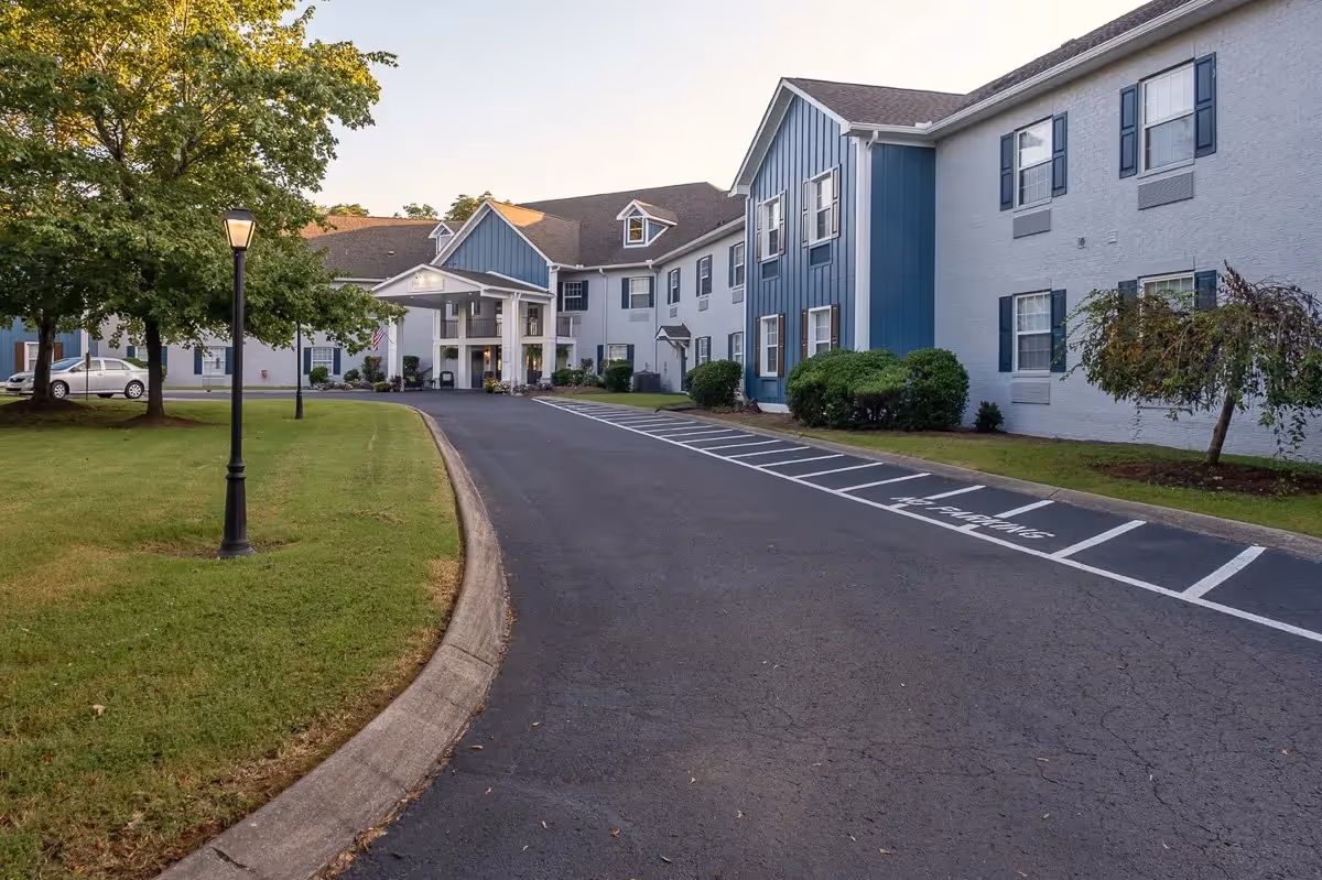 Exterior view of The Village at Bellevue Nashville Assisted Living facility showing a two-story building with blue and white siding, multiple windows with shutters, a covered entrance, a driveway, parking spaces, green lawn, trees, and a lamppost.
