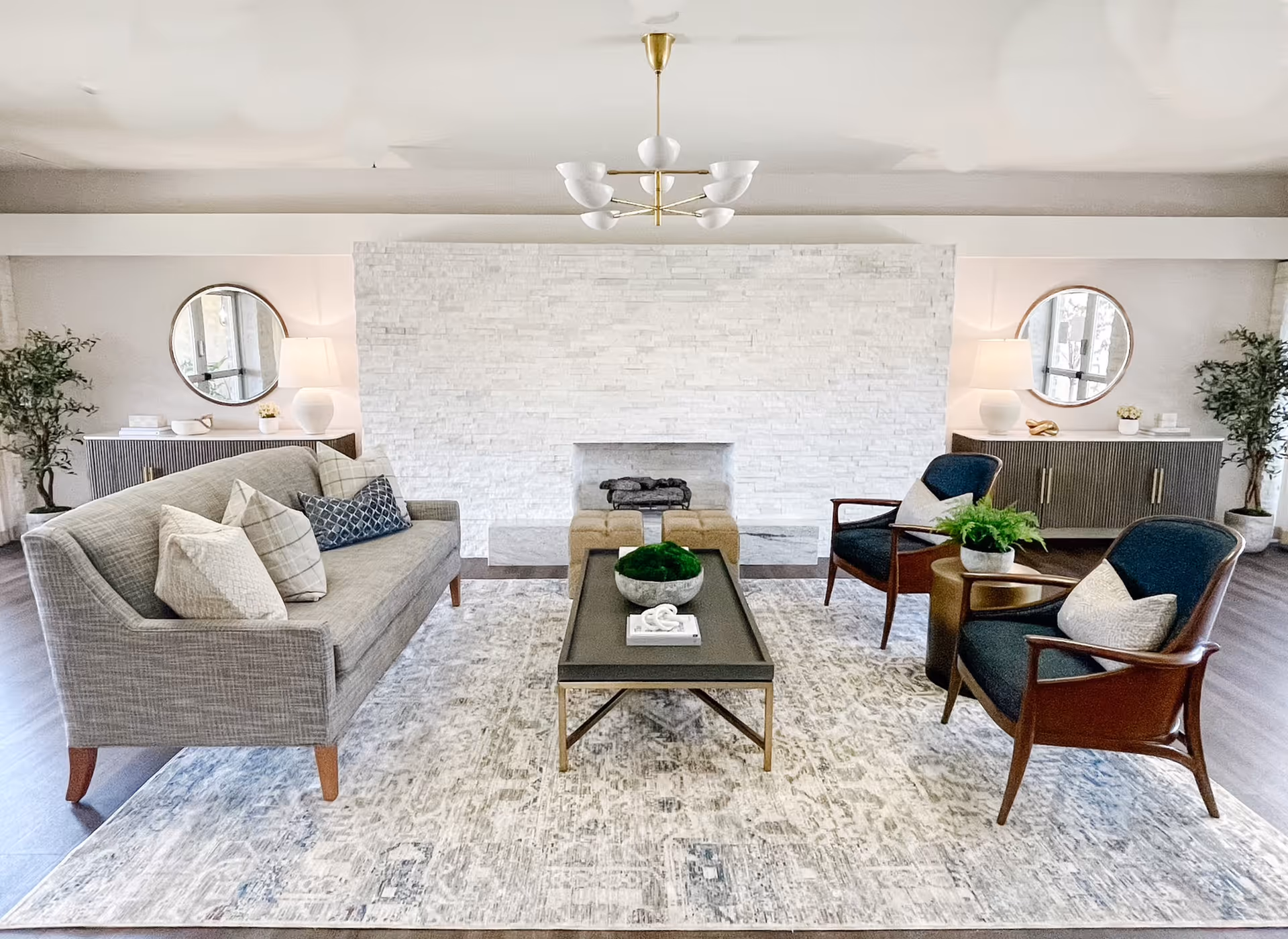 A bright and modern living room with a gray sofa adorned with multiple pillows, two dark blue armchairs with light-colored cushions, and a rectangular coffee table with decorative items. The room features a white textured stone fireplace in the center, two round mirrors above sideboards on either side of the fireplace, table lamps, and potted plants. A patterned area rug covers the wooden floor.