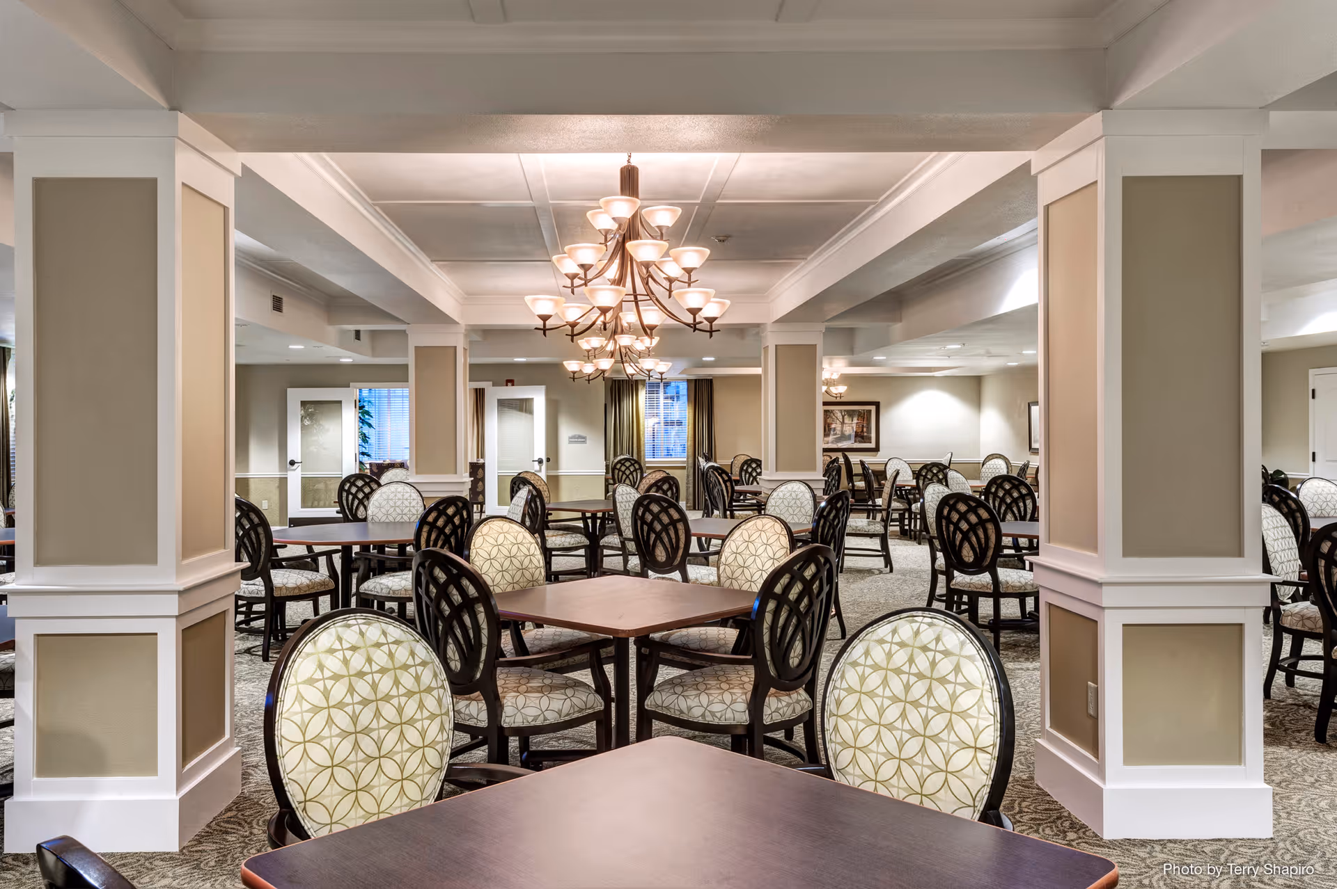 A spacious dining room with multiple square tables and cushioned chairs featuring patterned upholstery. The room has beige walls with white trim, decorative columns, and elegant chandeliers hanging from the ceiling. There are windows with curtains and framed artwork on the walls.