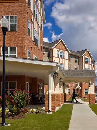 Exterior view of a multi-story senior living facility with brick and beige siding, featuring a covered entrance with columns, a sidewalk, green lawn, and a person walking near the entrance under a partly cloudy sky.