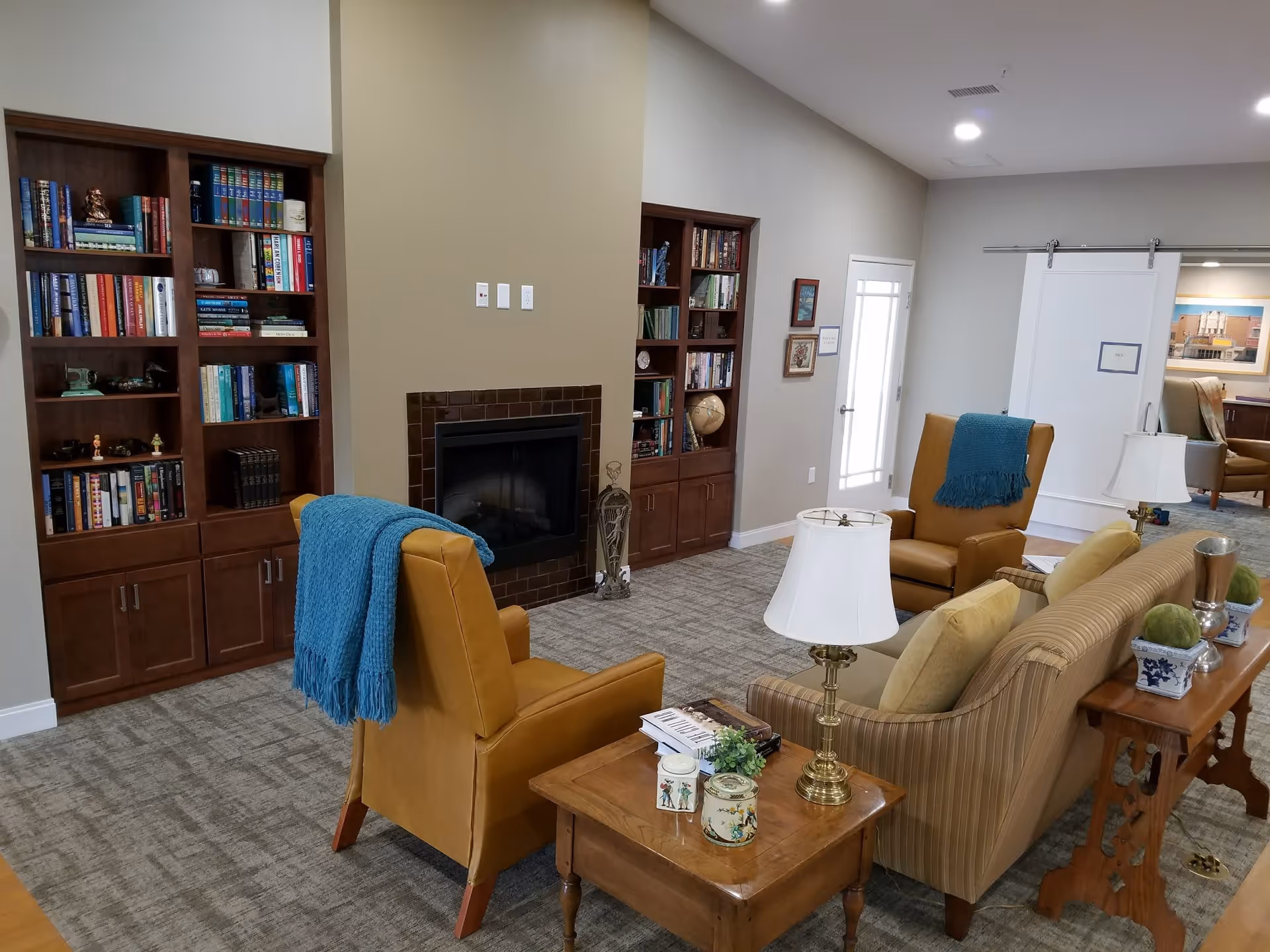A cozy living room area in a senior living facility featuring two mustard yellow armchairs with blue throws, a striped sofa with beige cushions, wooden side tables with lamps and decorative items, built-in wooden bookshelves filled with books and decor, and a fireplace with a brown tile surround. The room has neutral-colored walls and carpeted flooring, with a white sliding door leading to another room.