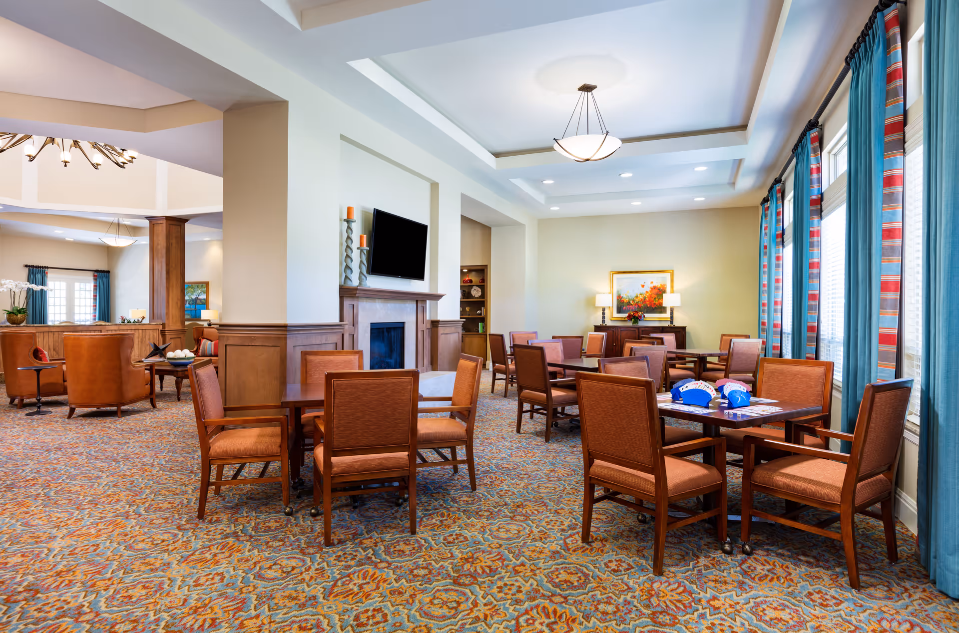 A spacious and well-lit common area in a senior living facility featuring multiple wooden tables with orange cushioned chairs arranged around them. The room has a patterned carpet, large windows with blue and red striped curtains, a fireplace with a mounted TV above it, and a sideboard with a colorful painting and lamps. The area appears comfortable and inviting for socializing or activities.