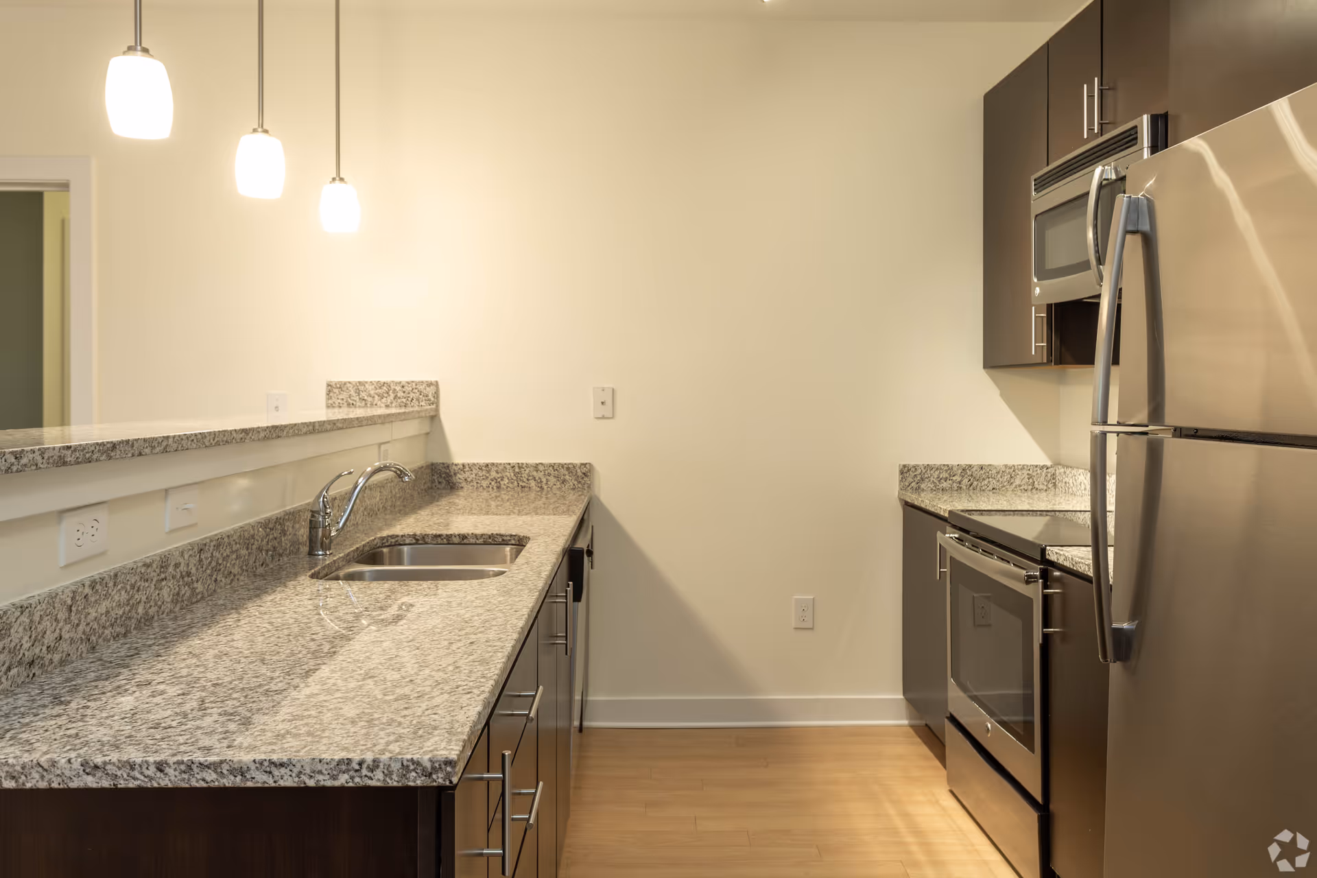 Modern kitchen with granite countertops, stainless steel refrigerator, oven, microwave, and dark wood cabinets. Three pendant lights hang from the ceiling above the counter with a built-in sink.