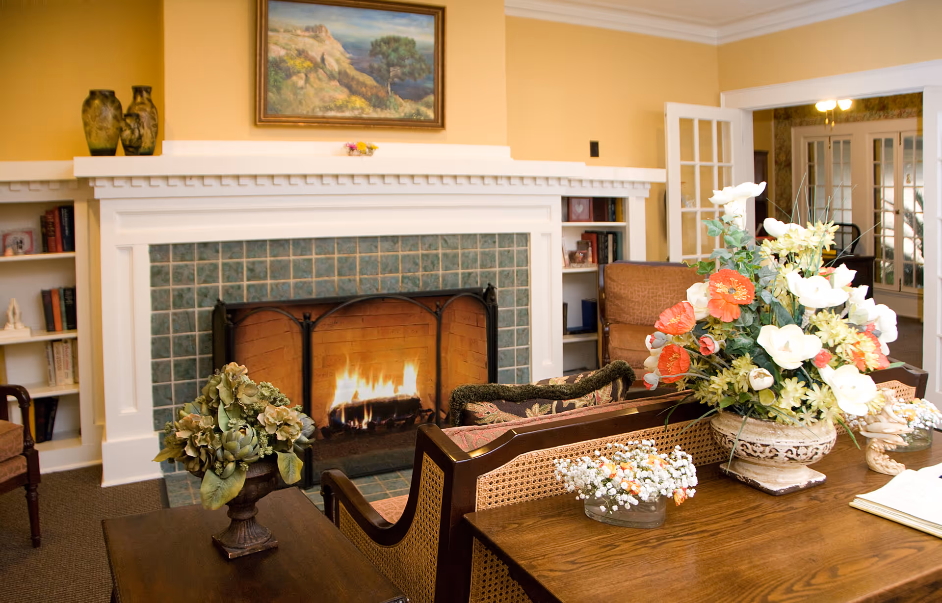 A cozy living room with a lit fireplace surrounded by green tiles and a white mantel. There are built-in bookshelves on either side of the fireplace with books and decorative items. A wooden table in the foreground holds a large floral arrangement with white and orange flowers, and smaller flower vases. The walls are painted yellow, and a framed landscape painting hangs above the fireplace. French doors lead to another room in the background.