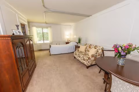 Carpeted private room with a single bed by a window, a floral loveseat, wooden armoire, and a round table with a vase of flowers.