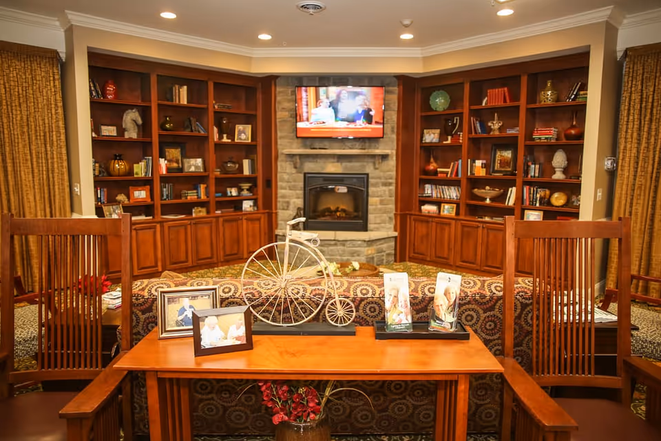 Community lounge with wooden bookshelves flanking a stone fireplace and wall-mounted TV, wooden chairs and a table with decorative items in the foreground.