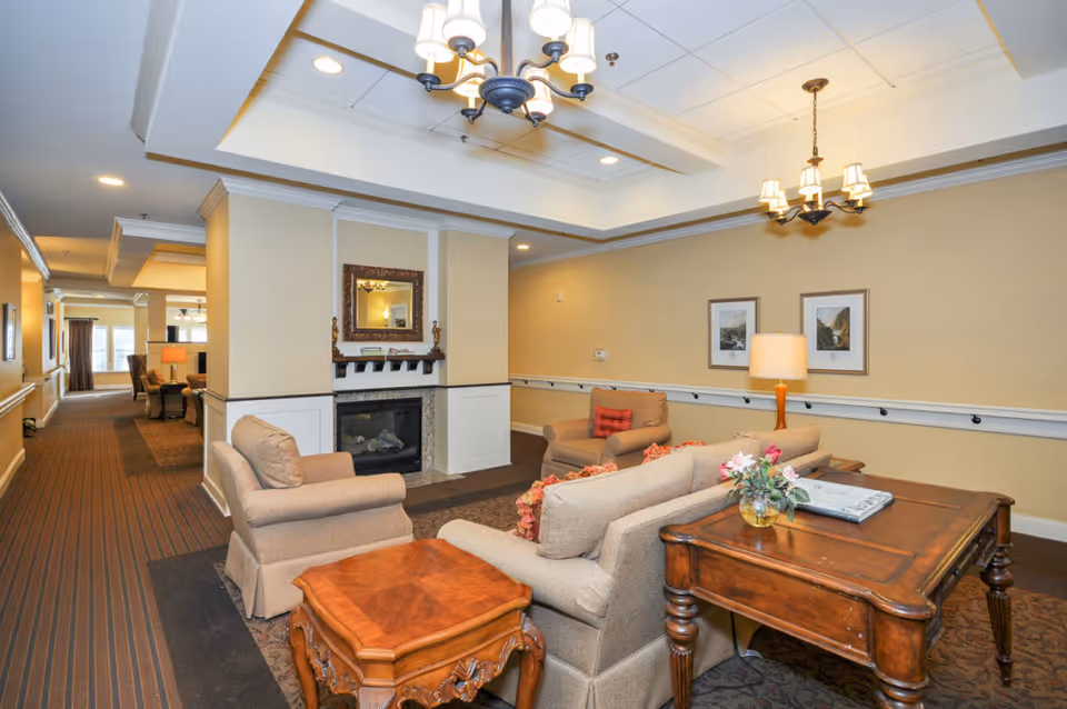 A cozy senior living facility common area with beige walls and carpeted floors. The space features comfortable beige armchairs and a sofa arranged around a fireplace with a decorative mirror above it. There are two wooden tables, one with a small flower arrangement and a book. The ceiling has recessed lighting and two chandeliers. The hallway extends to the left with more seating and windows at the end.