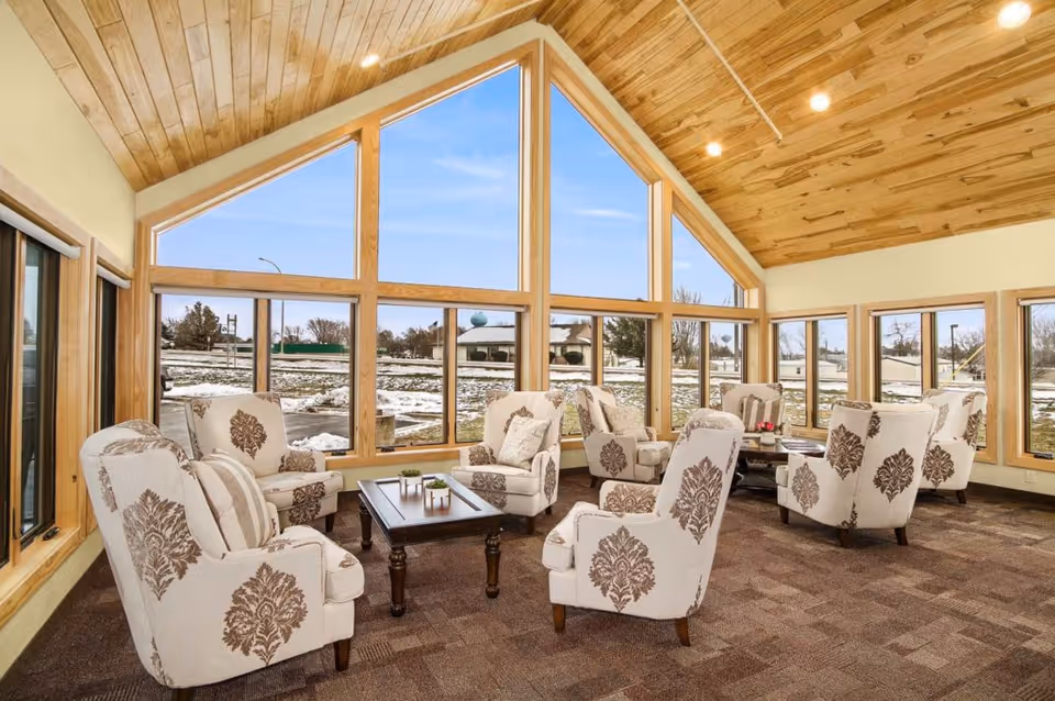 A bright and spacious living room with large triangular windows allowing natural light to fill the space. The room features multiple upholstered armchairs with a floral pattern arranged around two wooden coffee tables. The ceiling is made of wood panels with recessed lighting, and the floor is carpeted. Outside, there is a snowy landscape visible through the windows.