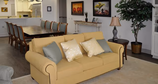 Living room with a yellow sofa and decorative pillows, side table and lamp, potted plant, and a dining table and kitchen visible in the background.