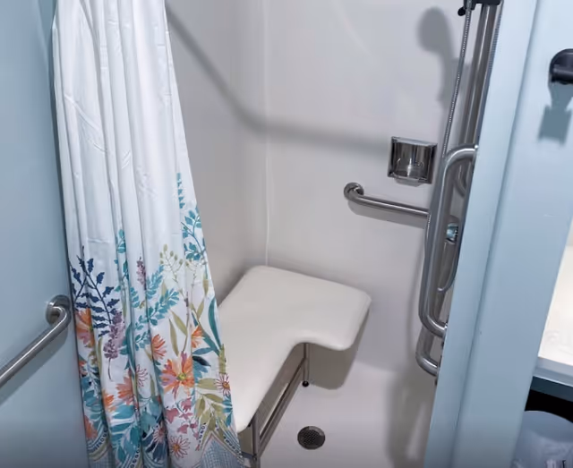 Shower area with a white foldable seat, grab bars, a handheld showerhead, and a floral-patterned shower curtain.