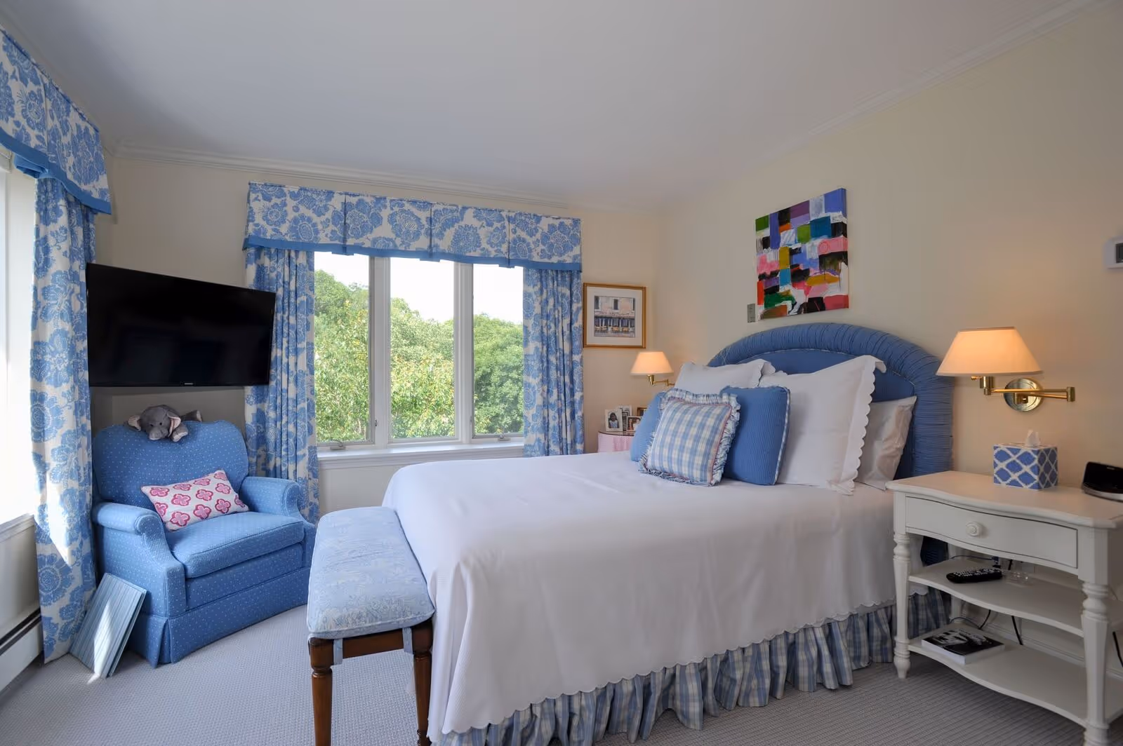 Bright bedroom with a made bed, blue upholstered chair, floral curtains at a large window, and bedside tables with lamps.