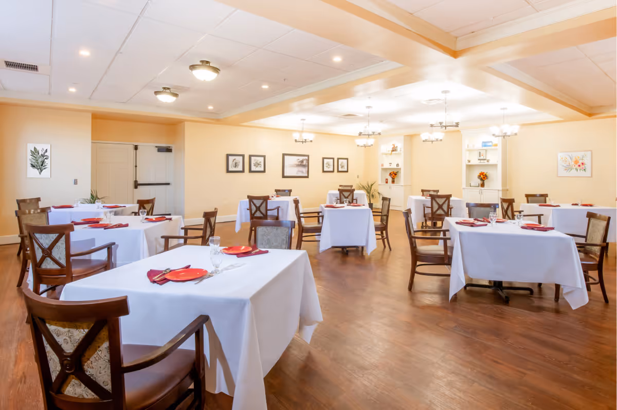 A dining room with several tables covered in white tablecloths, each set with red plates, napkins, and glassware. Wooden chairs with cushioned seats surround the tables. The room has warm beige walls adorned with framed botanical artwork and is illuminated by ceiling lights and chandeliers. The floor is wooden, and there are built-in shelves with decorative items and flowers.