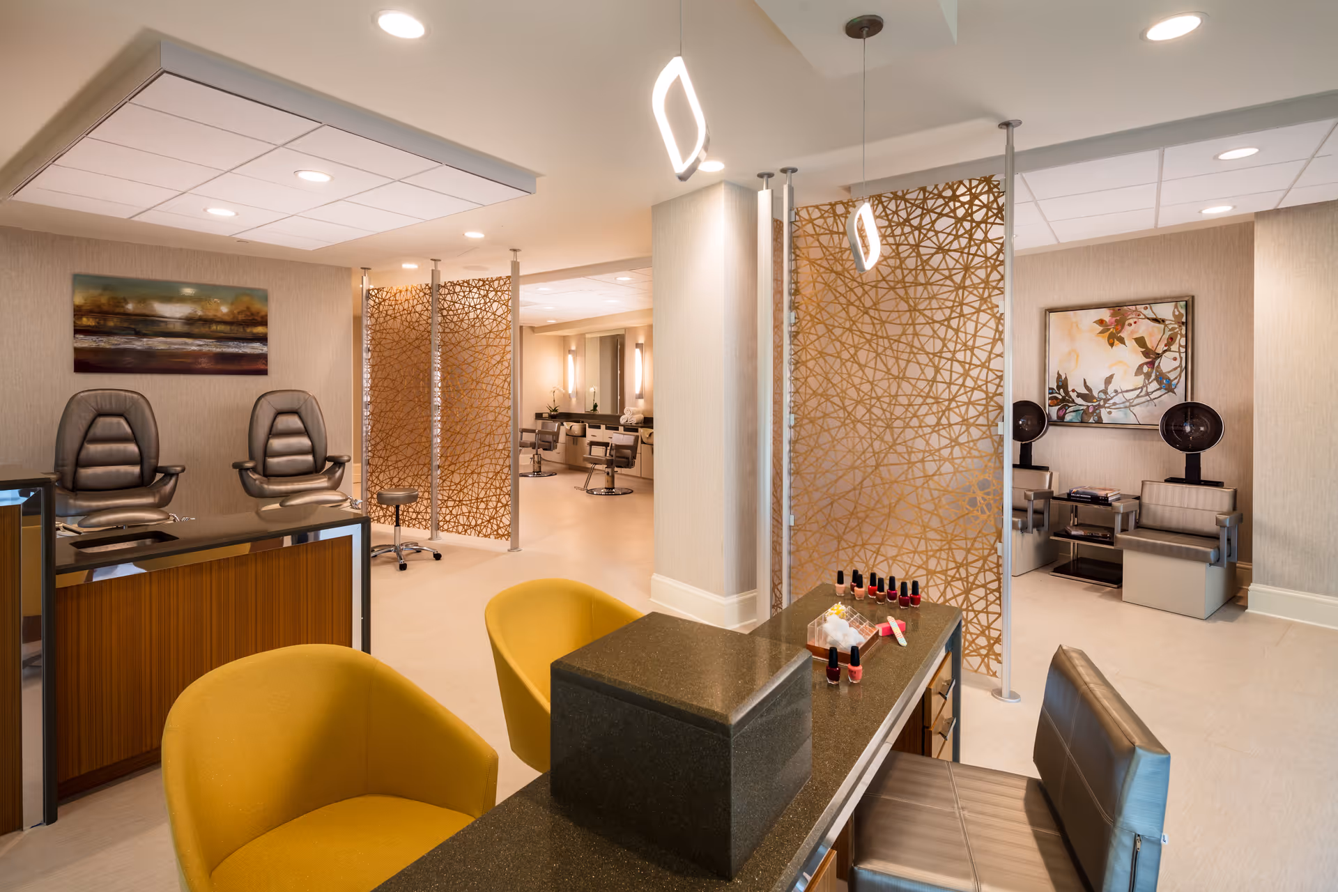 Interior view of a modern salon area in a senior living facility with yellow chairs at a manicure station displaying nail polish bottles, leather pedicure chairs, hair styling stations with mirrors and chairs, and decorative golden privacy screens.