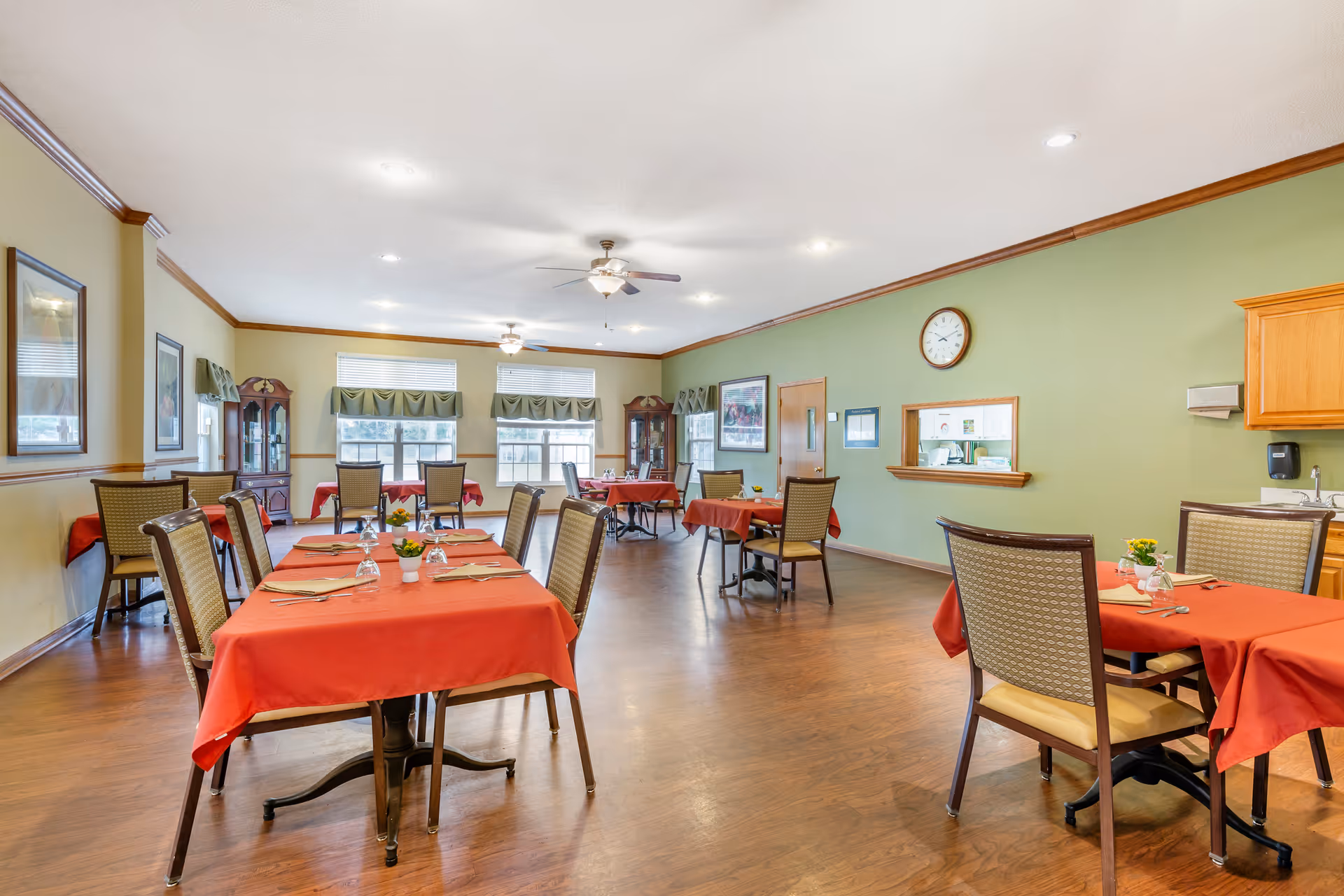 A dining room with several tables covered in red tablecloths, each set with napkins, utensils, and small flower pots. The room has wooden flooring, green and beige walls, ceiling fans, and large windows with green valances allowing natural light to enter. There are wooden cabinets and framed pictures on the walls.