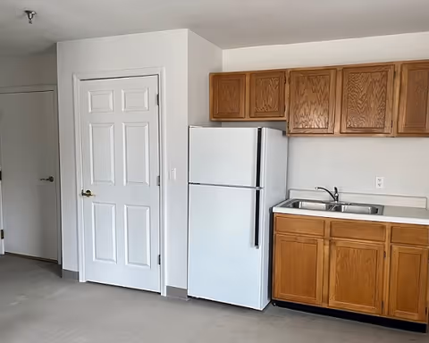 Small kitchenette with a white refrigerator, oak cabinets and a double sink beside a closed white door in a neutral room.