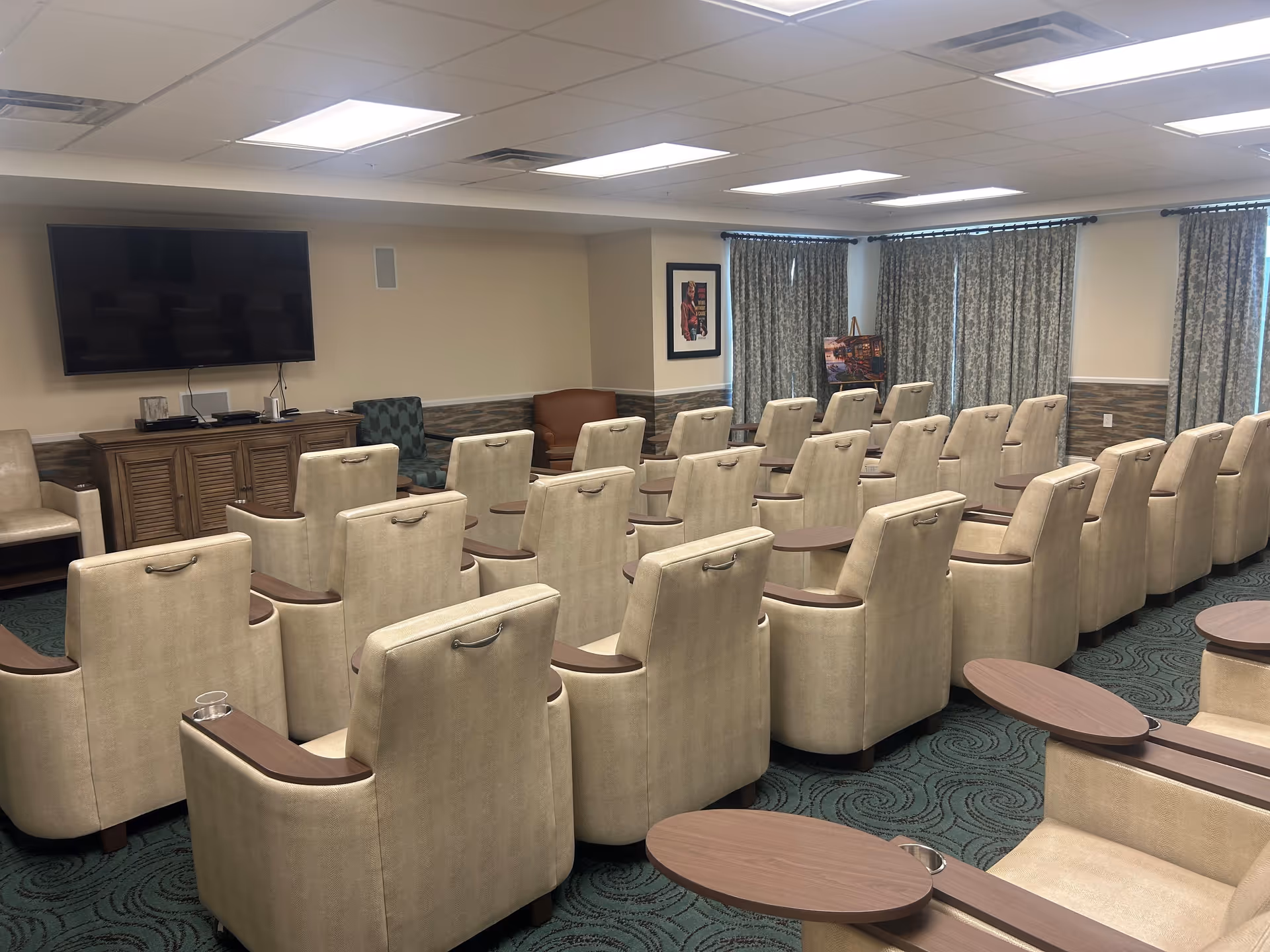 A senior living facility common room with multiple beige upholstered armchairs arranged in rows facing a large flat-screen TV mounted on the wall. The room has patterned green carpet, a wooden cabinet under the TV, and windows covered with patterned curtains. There are also a few additional chairs and framed artwork on the walls.