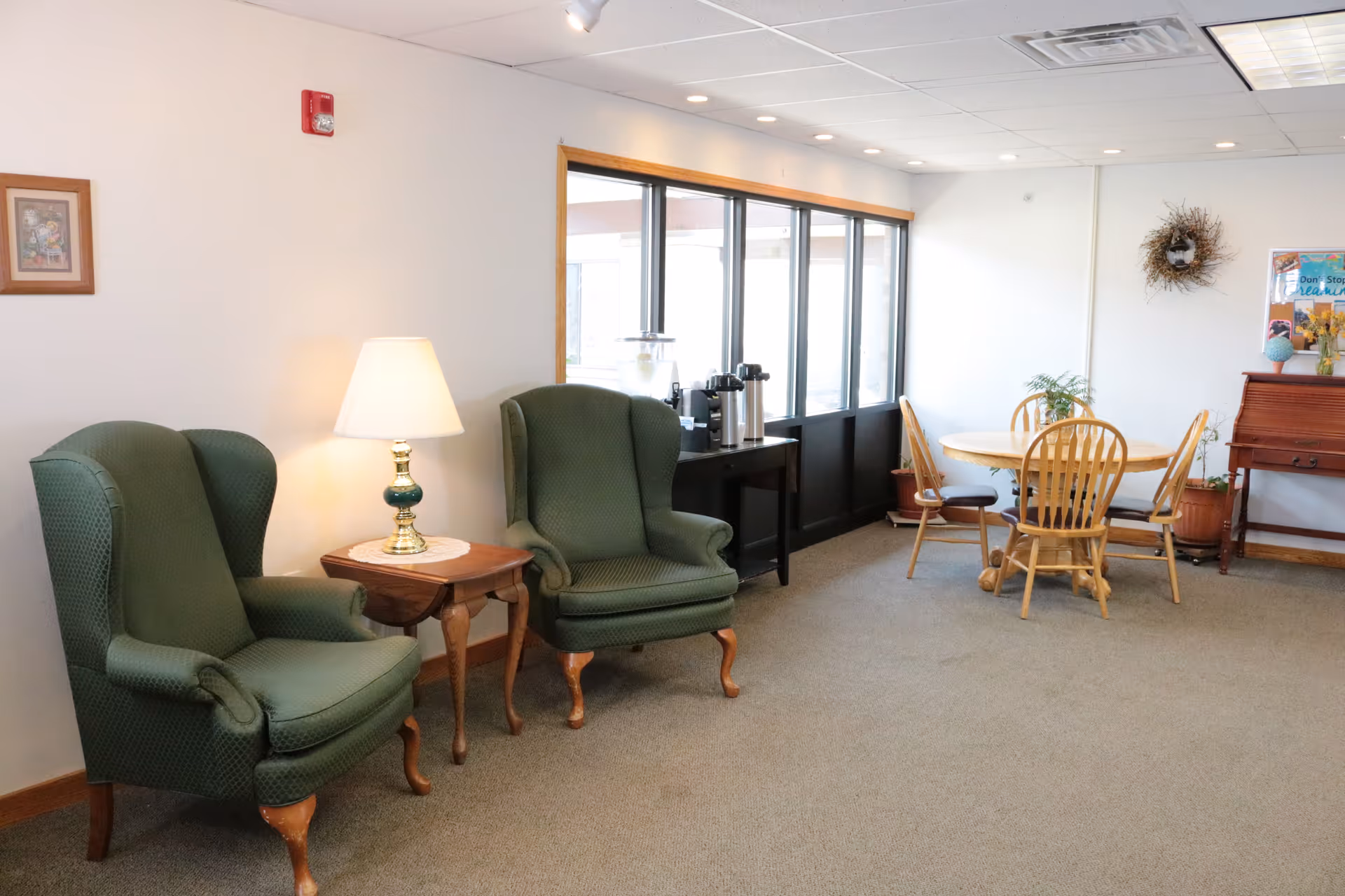 A cozy sitting area with two green upholstered armchairs separated by a wooden side table with a lamp. In the background, there is a round wooden table with four chairs, a small table with coffee and water dispensers, and a wall decorated with a wreath and framed pictures.