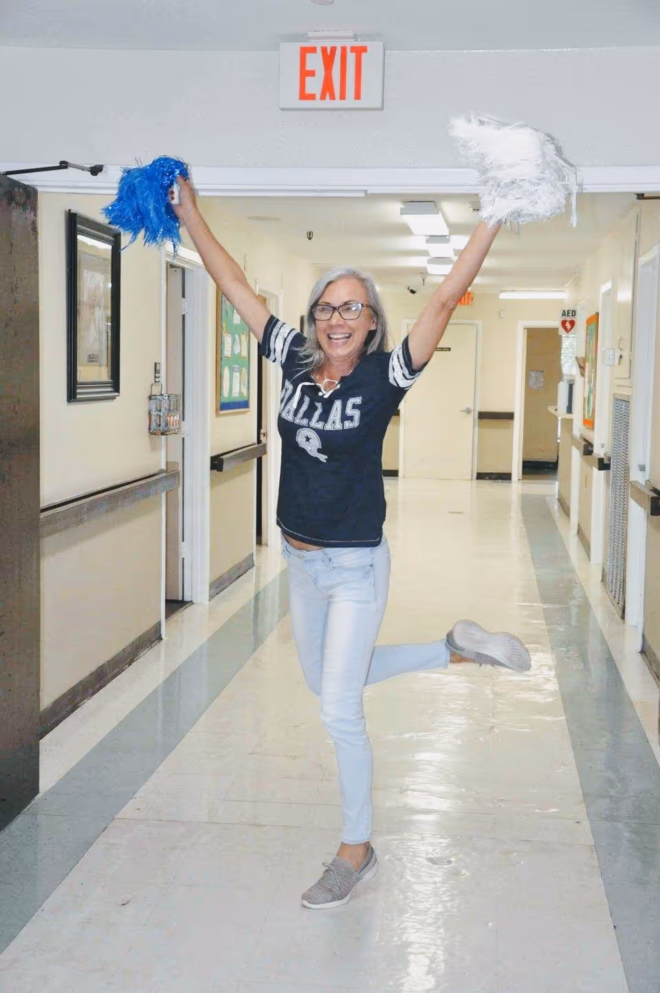 A woman wearing glasses, a Dallas Cowboys shirt, and light blue jeans is joyfully posing in a hallway with her arms raised, holding blue and white pom-poms. She is standing on one leg and smiling. The hallway has white walls, handrails, and an exit sign above the doorway behind her.