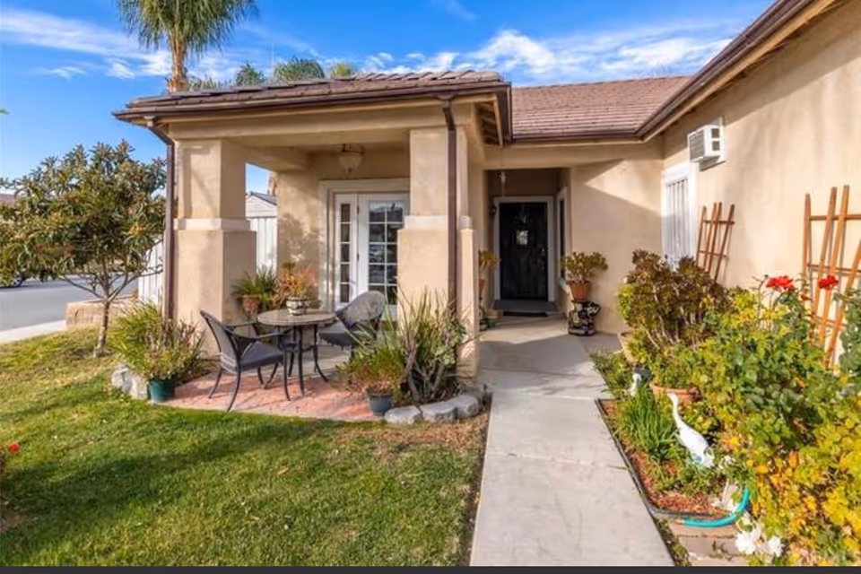 Front porch area of a residential building with a small patio set including a round table and two chairs. The porch is surrounded by plants and greenery, with a concrete walkway leading to the front door. The building has beige walls and a tiled roof under a partly cloudy sky.