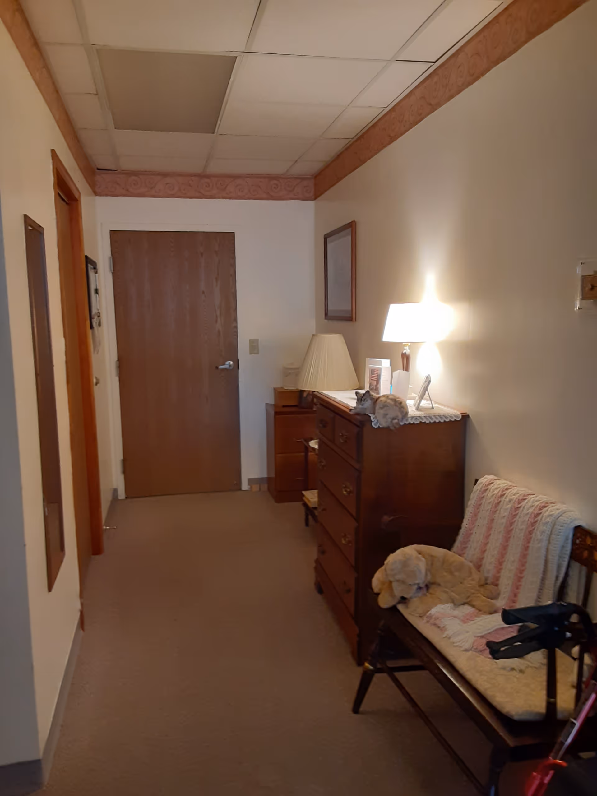 A hallway in a senior living facility with a wooden door at the end. On the right side, there is a wooden dresser with a lamp, framed picture, and decorative items on top. Next to the dresser is a wooden bench with a crocheted blanket, a stuffed dog, and a walker nearby. The walls are light-colored with a decorative border near the ceiling.