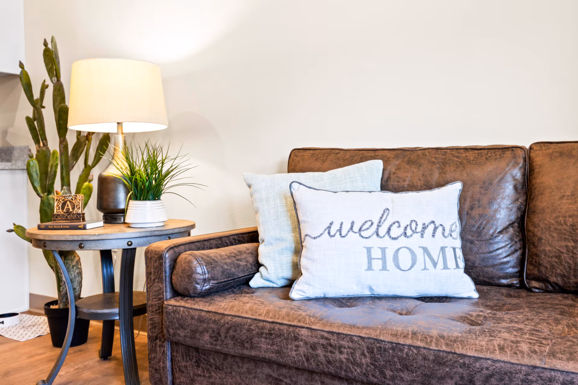Brown leather couch with a 'welcome HOME' pillow beside a round side table holding a lamp and potted plants.
