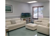 A bright senior living lounge with cream sofas, ottoman, aquarium, and blinds.