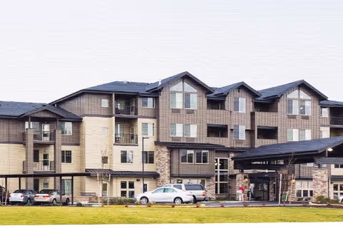 Exterior view of a multi-story senior living facility building with beige and brown siding, multiple windows, balconies, and a covered entrance. Several cars are parked in front of the building on a paved driveway, and a grassy lawn is visible in the foreground.