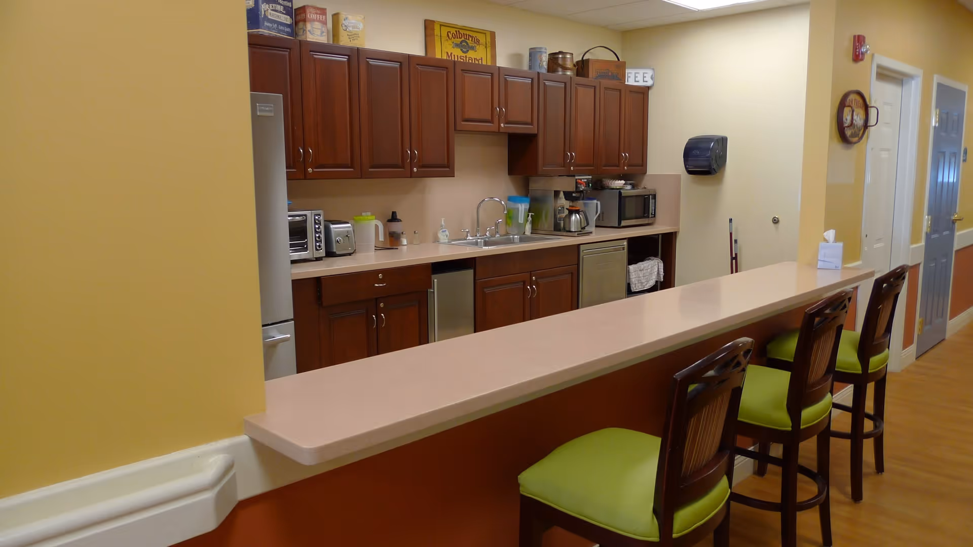 Communal kitchenette with a long counter and three green-cushioned bar stools in front of wooden cabinets, sink and small appliances.