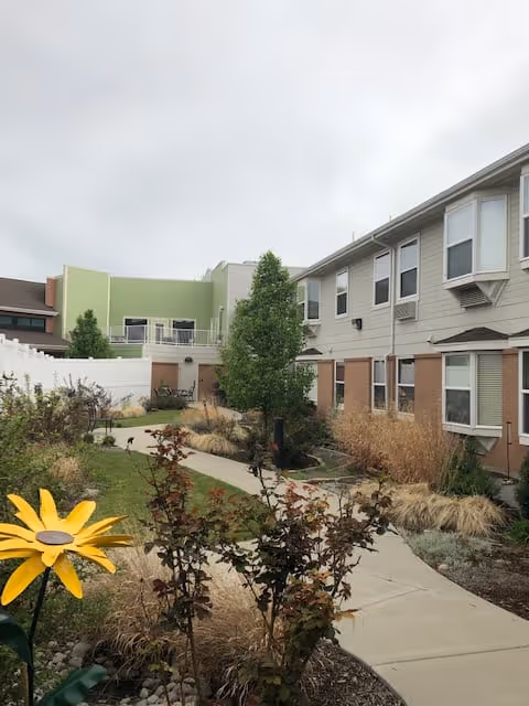 Outdoor garden area with a concrete pathway winding through landscaped plants and shrubs. There is a large yellow decorative flower in the foreground. The garden is bordered by a two-story building with multiple windows and a white fence on one side. The sky is overcast.