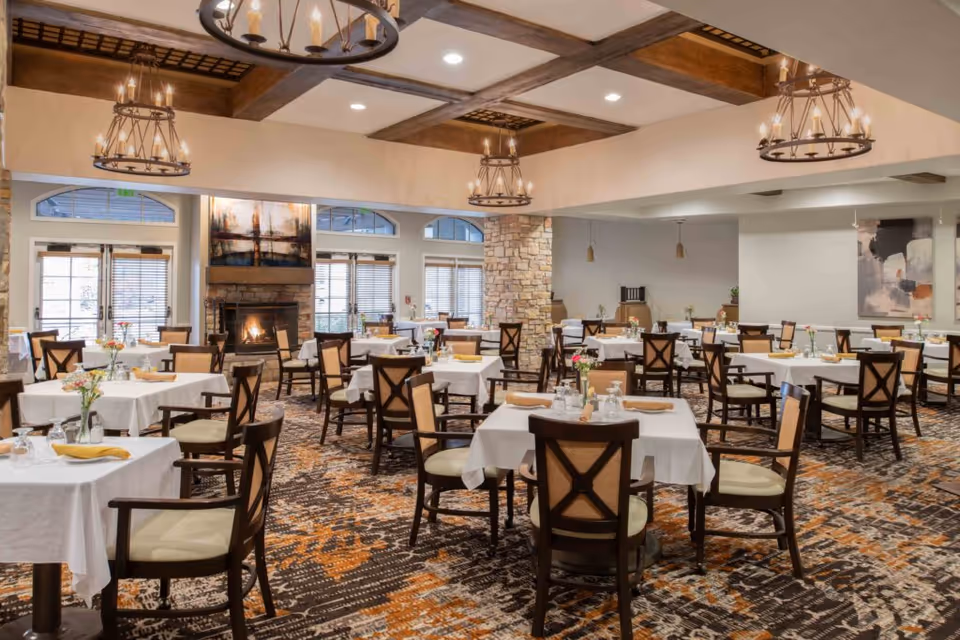 A spacious dining room with multiple tables covered in white tablecloths, each set with glassware, napkins, and small flower vases. The room features wooden chairs with cushioned seats, a patterned carpet in shades of brown and orange, a stone fireplace with a painting above it, large windows with blinds, and decorative chandeliers hanging from a ceiling with wooden beams.