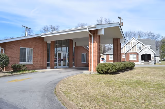 Exterior view of a brick building with a covered entrance supported by brick columns. There is a driveway leading up to the entrance and some bushes and grass around the building. Another house-like building is visible in the background under a clear sky.
