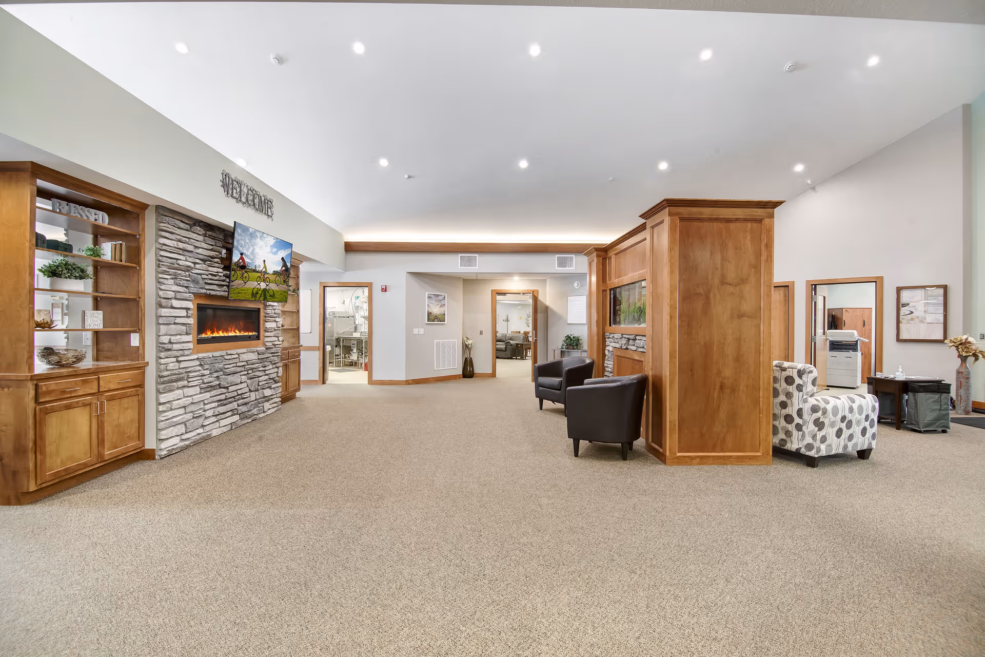 Spacious senior living facility common area with a stone fireplace, wall-mounted TV, wood cabinetry, and seating.