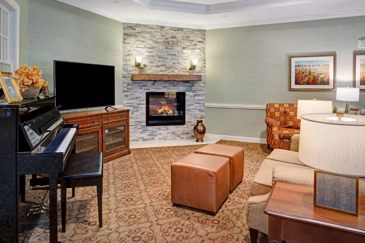 A cozy living room with a patterned carpet, a stone fireplace with a wooden mantle and two wall sconces, a flat-screen TV on a wooden cabinet, a black piano with a bench, two brown ottomans, a beige sofa, an orange patterned armchair, a wooden side table with a lamp, and framed artwork on the walls.