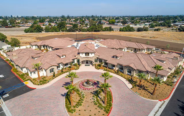 Aerial view of a single-story senior living facility with terracotta-tiled roofs, a circular driveway and central fountain surrounded by landscaping.