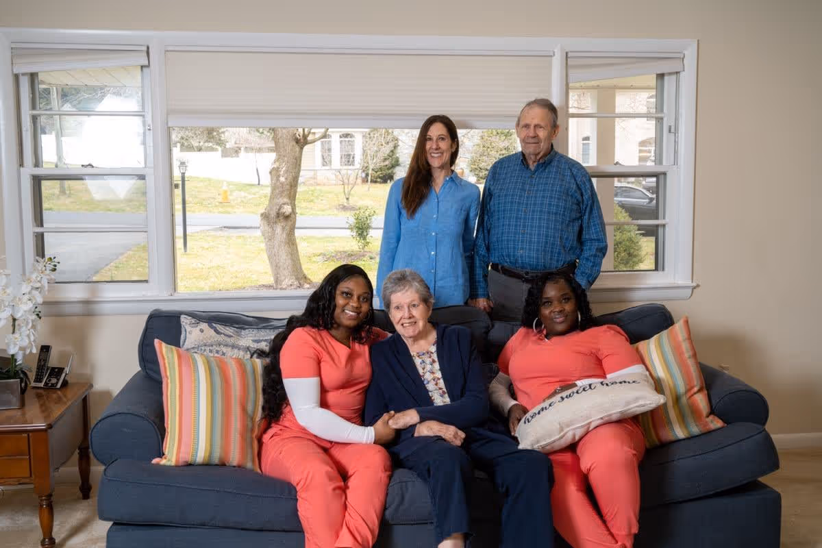 Five people in a living room setting with a large window behind them. Two elderly individuals, a man and a woman, stand behind a blue couch. Two women in matching coral scrubs sit on either side of an elderly woman seated in the middle of the couch. The couch has colorful striped pillows and a pillow with the words 'home sweet home'. A wooden side table with a phone and a white orchid plant is visible to the left.