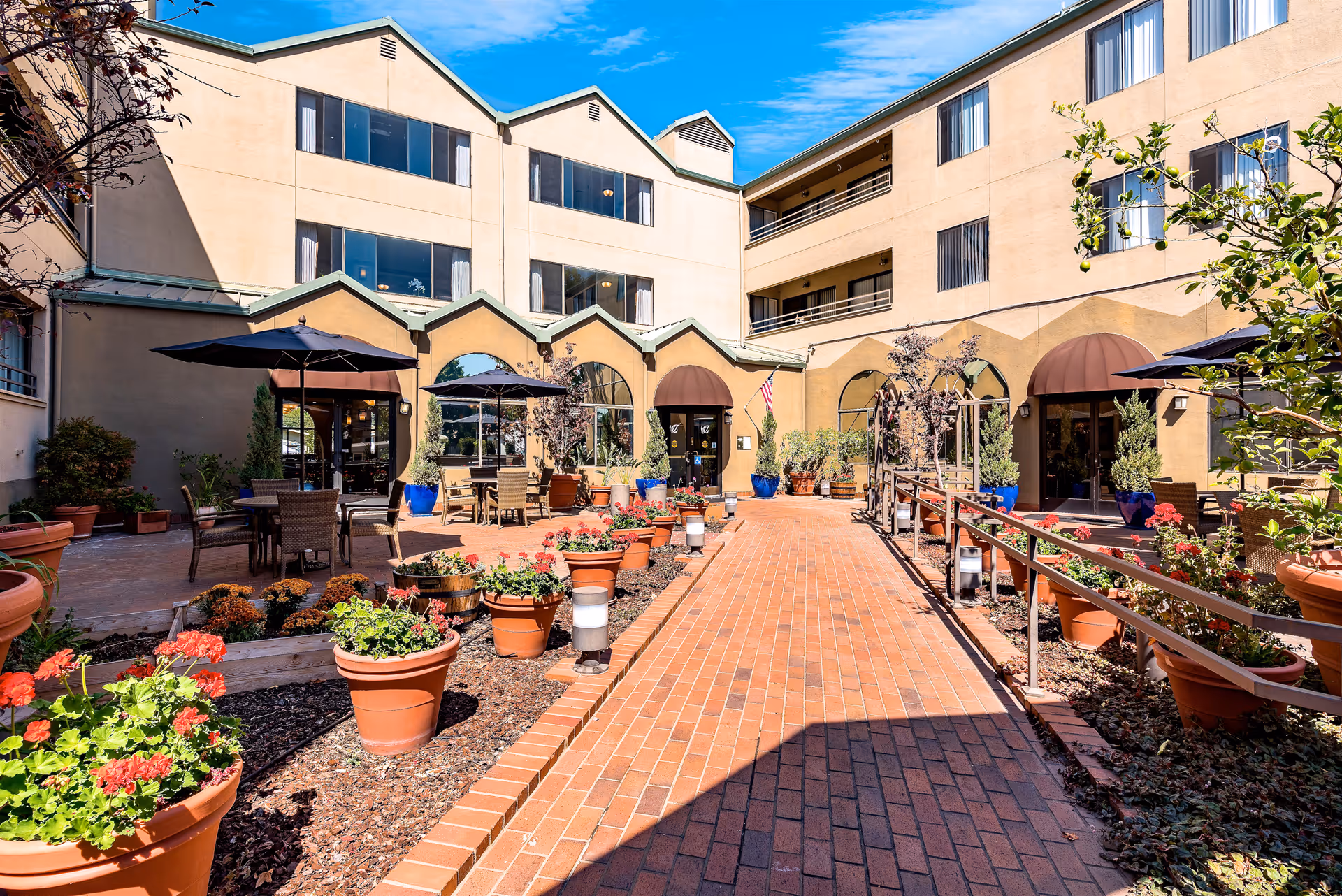 Sunny central courtyard with a brick walkway, potted flowers and patio seating surrounded by a three-story building.