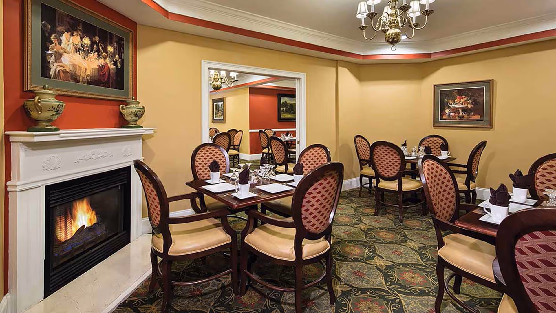 Cozy dining room with multiple set tables and upholstered chairs, a lit fireplace and chandelier.
