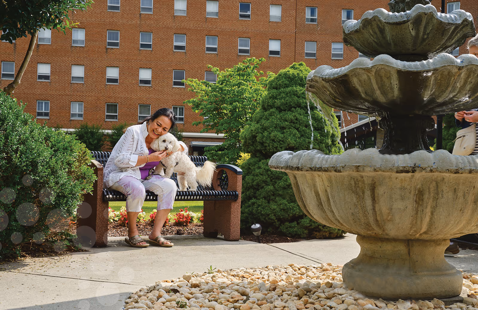 A woman sits on a bench in a courtyard petting a small white dog near a large stone fountain with a brick building in the background.