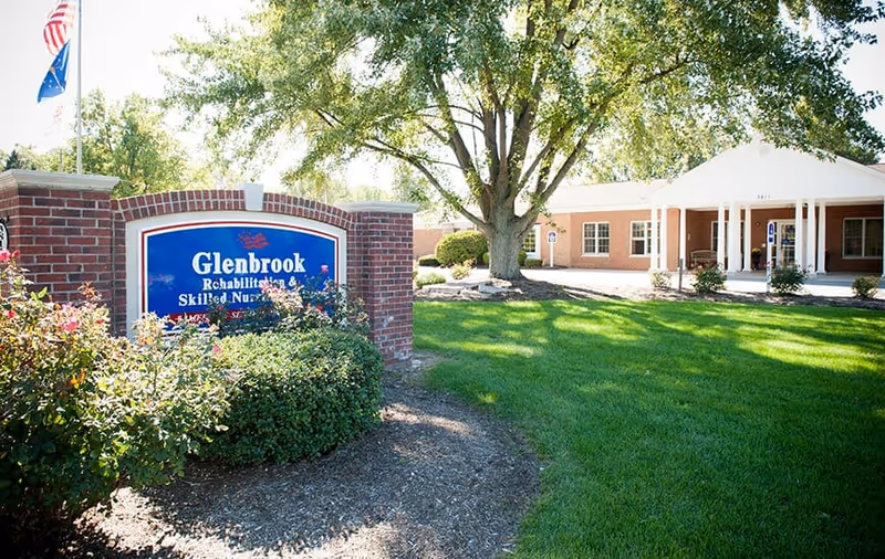 Exterior view of Glenbrook Rehabilitation and Skilled Nursing Center with a brick sign in the foreground surrounded by bushes and flowers, a large tree, and the building entrance visible in the background on a sunny day.