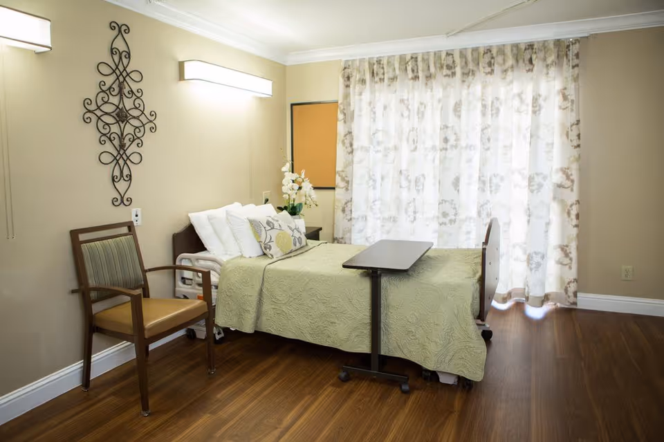 A tidy nursing facility bedroom with a single bed, overbed table, chair, decorative wall art and patterned curtains.