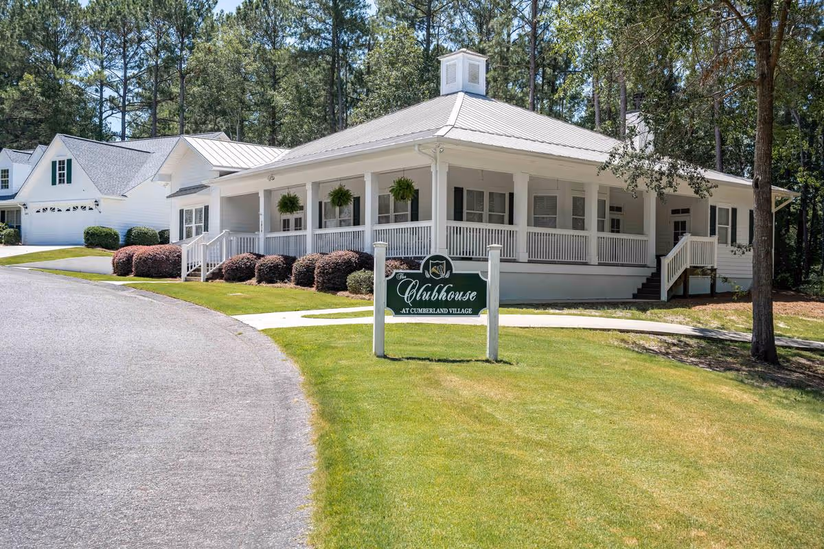 White single-story clubhouse with a wraparound porch, hanging ferns, and a green 'Clubhouse' sign on a manicured lawn.